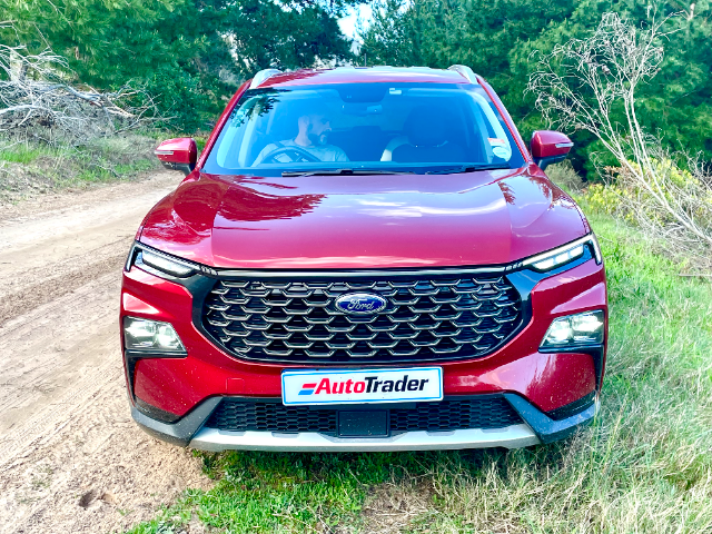 A Ruby Red Ford Territory parked on the grass next to a rural road with green trees in the background.