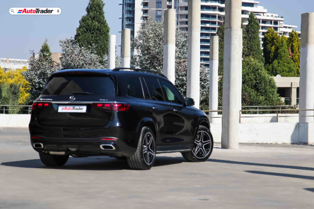 A black Mercedes-Benz GLS450d AMG Line parked in an outdoor parking area, featuring a sleek design with distinctive AMG styling. The vehicle is positioned to show its rear and side profile, highlighting the LED taillights and sporty alloy wheels. In the background, there are modern buildings and greenery under a clear blue sky.