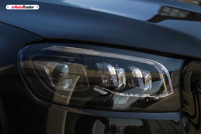 Close-up view of the headlight on a Mercedes-Benz GLS450d AMG Line. The image highlights the intricate design of the LED lights and the sleek, reflective surface of the car's dark blue body. The AutoTrader logo is visible in the top left corner.