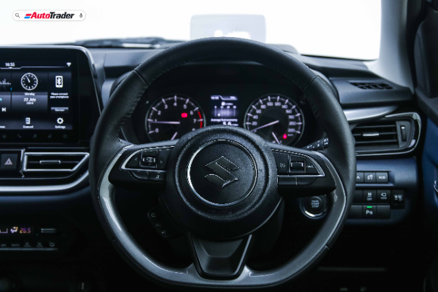 The image shows the interior of a Suzuki Baleno, focusing on the steering wheel and dashboard. The steering wheel features the Suzuki logo in the center and various control buttons on the spokes. Behind the steering wheel, the instrument cluster displays the speedometer, tachometer, and other gauges. To the left, the infotainment system screen is visible, showing various settings and a clock. The dashboard also includes air vents, climate control buttons, and other vehicle controls. The overall design is modern and functional. :+1: 1 :-1: 1