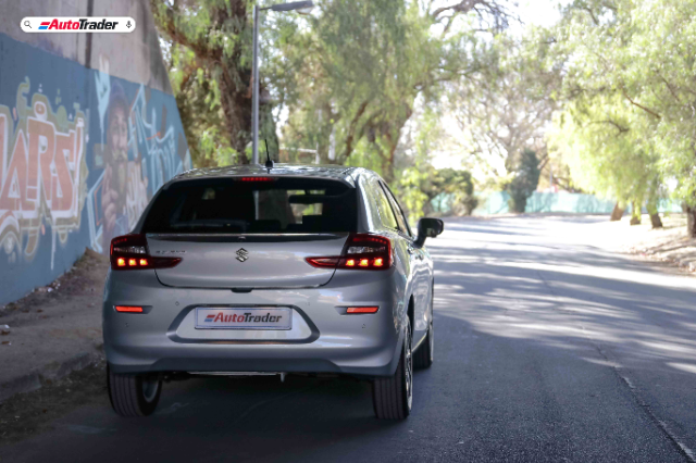 A silver Suzuki Baleno is shown from the rear, parked on a shaded road with trees on both sides. The car has its taillights on, and a license plate with the "AutoTrader" logo is visible. To the left, there is a wall with colorful graffiti art. The scene is well-lit with natural light filtering through the trees.