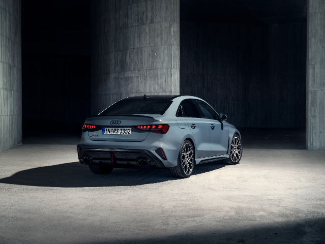 A sleek, grey Audi RS3 saloon is parked in a dimly lit, industrial concrete structure. The car is viewed from the rear three-quarter angle, showcasing its aggressive rear diffuser, quad exhaust tips, and distinctive LED taillights. The license plate reads "IN RS 3352." The car's sporty design is accentuated by its large alloy wheels and low stance.