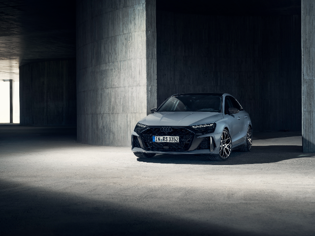 A sleek, gray Audi RS3 saloon is parked in a dimly lit, modern concrete structure. The car's aggressive front grille and sharp LED headlights are prominently displayed, highlighting its sporty design. The setting emphasizes the car's sophisticated and powerful presence.