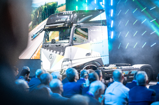 A futuristic Mercedes-Benz truck is showcased on stage in front of an audience, with a large screen displaying the truck in action on a highway.