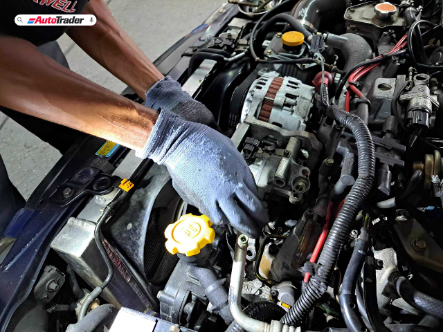 A mechanic wearing gloves is working on the engine of a Subaru Impreza, replacing the air conditioning compressor at Bens Auto Aircon in Randburg. The image shows the engine bay with various components and the mechanic's hands focused on the compressor. The AutoTrader logo is visible in the top left corner.