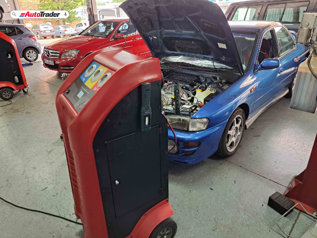 A blue Subaru Impreza with its hood open is being serviced at Bens Auto Aircon in Randburg. The air conditioning compressor is being replaced, and a red and black air conditioning service machine is positioned in front of the car. Other vehicles, including a red Mercedes-Benz, are visible in the background inside the garage.