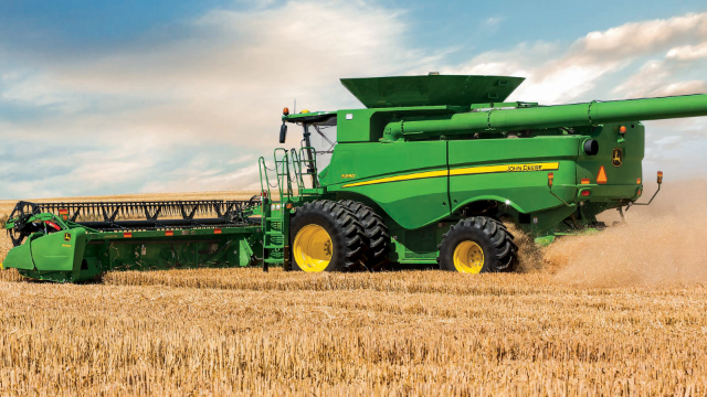A green John Deere combine harvester is working in a golden wheat field under a partly cloudy sky.