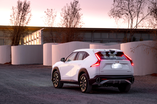 A white GAC Emkoo SUV is parked outdoors during sunset. The car is facing away, showcasing its sleek rear design and distinctive taillights. The background features modern architectural elements and sparse trees, adding to the serene and contemporary setting.