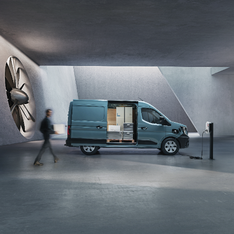 A blue delivery van with its side door open, revealing packages inside, is parked in a modern, industrial setting with a person walking by and the van connected to a charging station.