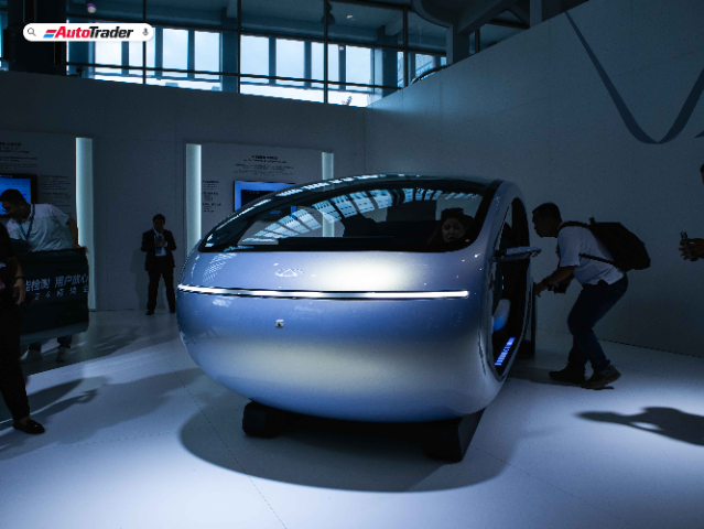 A futuristic, pod-like vehicle is displayed at the 2024 Chery International User Summit. The vehicle has a sleek, rounded design with a smooth, metallic blue exterior and a single horizontal light strip on the front. Several people are gathered around the vehicle, with one person inside and others examining it closely. The setting appears to be an indoor exhibition space with large windows and informational posters on the walls. The AutoTrader logo is visible in the top left corner of the image.