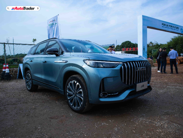 A blue Jaecoo J8 SUV is parked on a dirt road at an outdoor event. The vehicle features a prominent front grille with vertical slats and sleek, modern headlights. The car is surrounded by a few people in the background, and there is a banner and an archway with the text "FOR US, FOR FUTURE" visible. The AutoTrader logo is displayed in the top left corner of the image.
