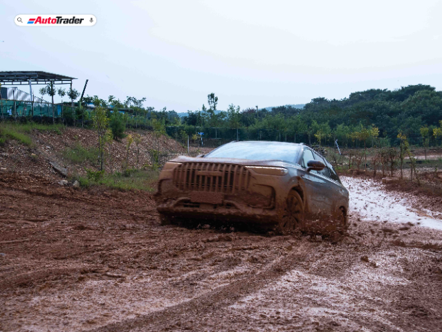 A Jaecoo J8 SUV is driving through a muddy off-road track, splashing mud around. The vehicle is covered in mud, indicating it is in the midst of an off-road adventure. The background shows a rural area with trees and a small structure on the left side. The image has the AutoTrader logo in the top left corner.