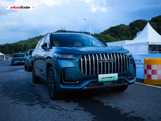 A Jaecoo J8 PHEV (Plug-in Hybrid Electric Vehicle) is prominently displayed in the foreground of the image. The vehicle has a sleek, modern design with a shiny blue exterior and a distinctive front grille featuring vertical chrome bars and the Jaecoo logo. The car is parked on a wet road, with another vehicle visible behind it. In the background, there is a white tent and some greenery, indicating an outdoor event or exhibition. The AutoTrader logo is visible in the top left corner of the image.