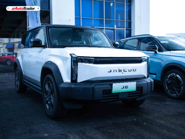 A white Jaecoo J6 SUV is parked in front of a building with large blue windows. The vehicle has a modern, boxy design with black accents and a prominent front grille displaying the "Jaecoo" logo. The license plate reads "J6." Another blue SUV is parked next to it. The image also features an "AutoTrader" logo in the top left corner.