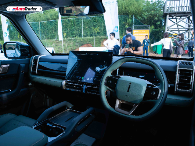 The image shows the interior of the Jaecoo J6, featuring a modern and sleek design. The dashboard includes a large central touchscreen display with various controls and information. The steering wheel has a minimalist design with integrated buttons. The interior is equipped with comfortable seats and a spacious layout. The view through the windshield shows an outdoor event with people and banners in the background.