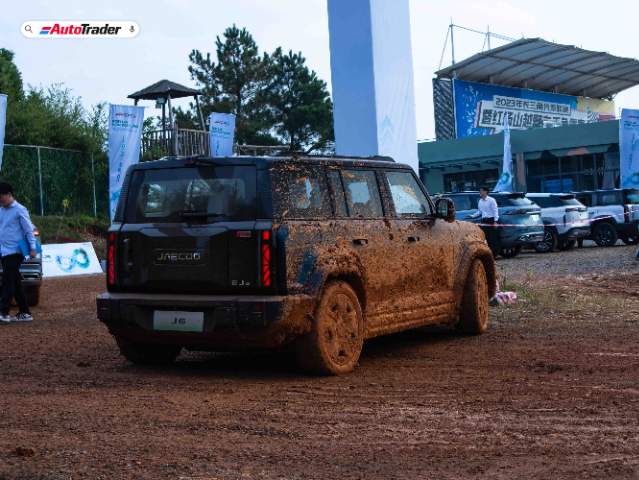 A Jaecoo J6 SUV is shown from the rear, covered in mud, indicating it has been driven off-road. The vehicle is parked on a dirt surface with a few people and other cars in the background. Banners and a building with a sign in Chinese are visible, suggesting an event or exhibition setting. The AutoTrader logo is present in the top left corner of the image.