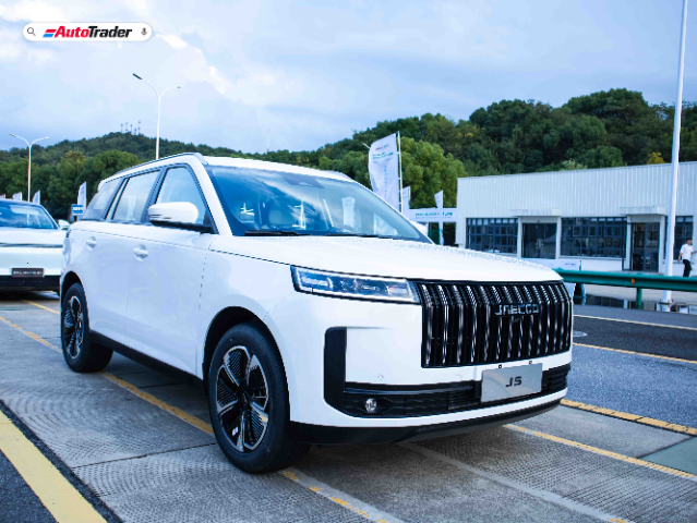 A white Jaecoo J5 SUV is parked on a paved area with a backdrop of greenery and a modern building. The vehicle features a sleek design with a prominent front grille, stylish headlights, and black alloy wheels. The AutoTrader logo is visible in the top left corner of the image.