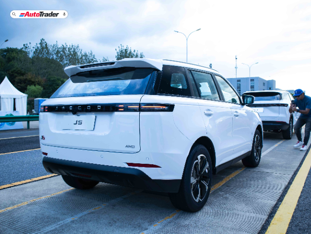 A white Jaecoo J5 SUV is parked on a road with a person taking a photo of it from the side. The vehicle has a sleek design with "JAECOO" written across the back and "J5" and "AWD" badges on the rear. The sky is overcast, and there are trees and a white tent in the background. The image has an "AutoTrader" logo in the top left corner.