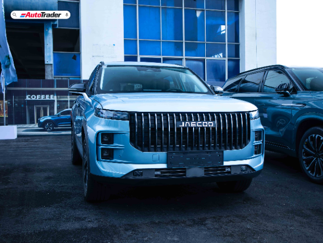 A front view of a blue Jaecoo J7 PHEV parked in front of a building with large windows. The vehicle features a prominent grille with vertical slats and sleek headlights. The AutoTrader logo is visible in the top left corner of the image. Another vehicle is partially visible on the right side.