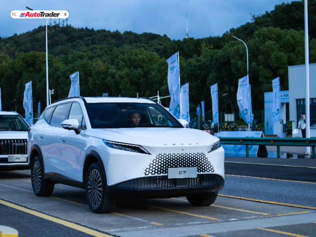 A white Omoda C7 SUV is parked on a road with a driver inside. The vehicle features a sleek, modern design with a distinctive front grille and stylish headlights. In the background, there are trees, flags, and a building, indicating an outdoor event or exhibition. The AutoTrader logo is visible in the top left corner of the image.