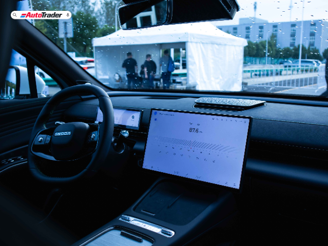 The image shows the interior of an Omoda C7 vehicle. The focus is on the driver's seat area, featuring a modern steering wheel with the Omoda logo prominently displayed. The dashboard includes a digital display behind the steering wheel and a large central touchscreen displaying a radio interface tuned to 87.6 MHz. The view through the windshield shows a rainy day with droplets on the glass and a white tent with people standing outside in the background. The overall ambiance is sleek and high-tech, emphasizing the vehicle's advanced features.
