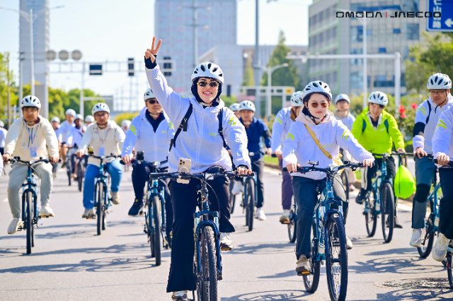 A group of cyclists, all wearing helmets and white jackets, participate in the Chery International User Summit IUCN cycling event. The cyclist in the foreground is smiling and making a peace sign with their hand. The event takes place on a sunny day in an urban area with buildings and traffic signs visible in the background.