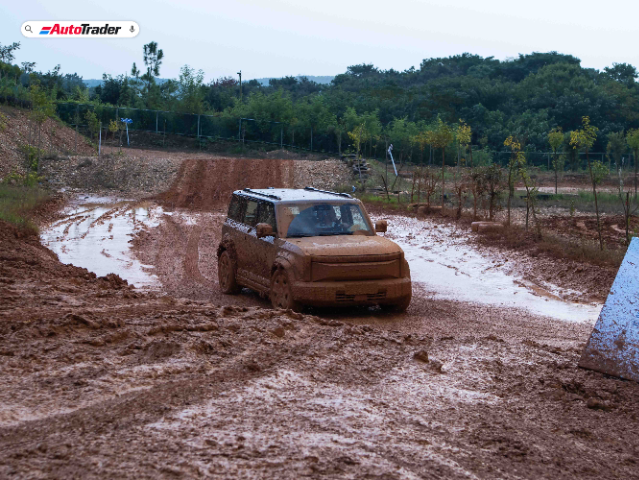 A Jaecoo J6 SUV is driving through a muddy off-road track, covered in mud. The vehicle is navigating a wet and rugged terrain surrounded by greenery and trees in the background. The AutoTrader logo is visible in the top left corner of the image.