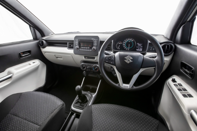 The image shows the interior of a Suzuki Ignis. The view is from the driver's seat, showcasing the dashboard, steering wheel, and front seats. The dashboard features a central infotainment system, air conditioning controls, and a speedometer. The steering wheel has the Suzuki logo and various control buttons. The interior design is a combination of black and light grey colors, with fabric seats and a manual gear shift. The driver's door panel includes window control buttons.