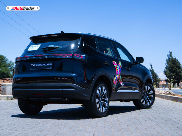 A black Chery Tiggo Cross SUV is parked on a paved surface under a clear blue sky. The vehicle's rear and right side are visible, showcasing its sleek design and modern taillights. A colorful, abstract decal is applied to the rear door. The AutoTrader logo is displayed in the top left corner of the image.