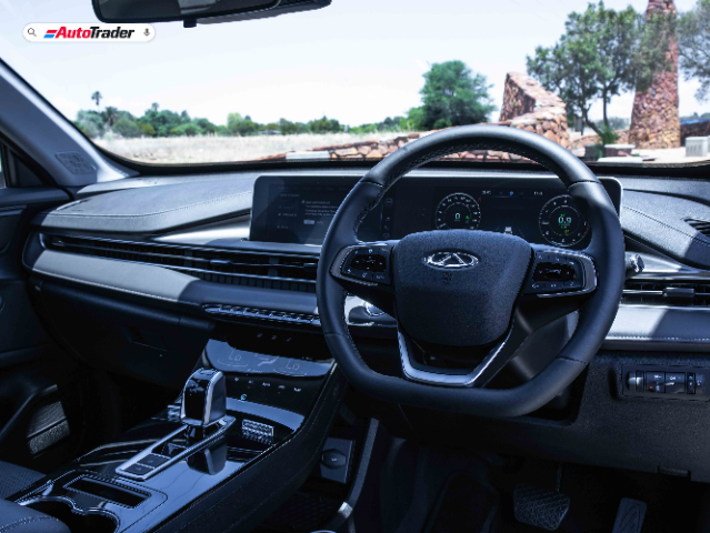 The image shows the interior of a Chery Tiggo Cross vehicle. The focus is on the driver's seat area, featuring a modern steering wheel with the Chery logo at the center. The dashboard is sleek and equipped with a digital display behind the steering wheel, showing various driving metrics. The center console includes a gear shifter and several control buttons. The interior design is sophisticated, with a combination of black and metallic accents. The windshield offers a view of an outdoor setting with trees and stone structures in the background. The AutoTrader logo is visible in the top left corner of the image.