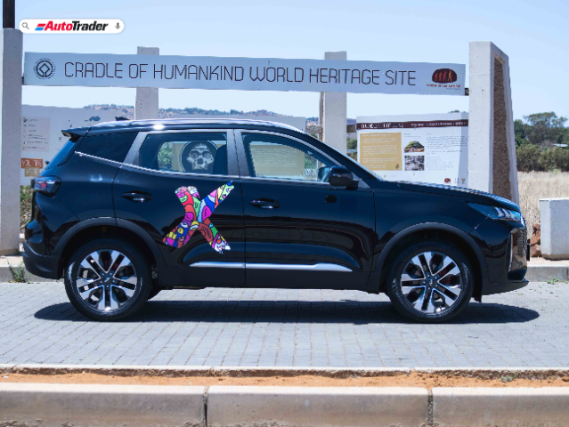 A black Chery Tiggo Cross SUV is parked on a paved area in front of a sign that reads "Cradle of Humankind World Heritage Site." The vehicle features a colorful, abstract "X" design on its side. The background includes informational plaques and a natural landscape. The image is branded with the AutoTrader logo in the top left corner.