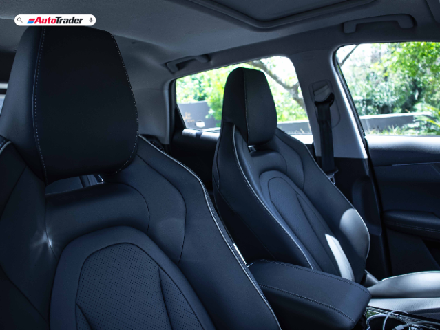 The image shows the interior of a Chery Tiggo Cross, focusing on the front seats. The seats are upholstered in black leather with white stitching, and the design appears modern and ergonomic. The image is taken from the back seat, providing a clear view of the headrests, seat contours, and part of the center console. The background shows a glimpse of greenery outside the car, indicating that the vehicle is parked in a well-lit area. The AutoTrader logo is visible in the top left corner of the image.