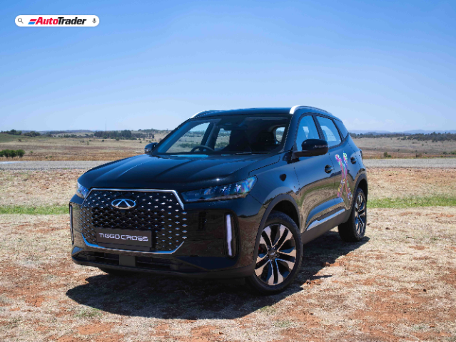A black Chery Tiggo Cross SUV is parked on a dry, grassy field with a clear blue sky in the background. The vehicle features a prominent front grille with a diamond pattern and sleek, modern headlights. The AutoTrader logo is visible in the top left corner of the image.