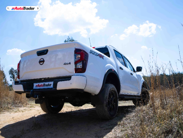 A white Nissan Navara PRO-4X pickup truck is shown from the rear, slightly elevated on a dirt path with dry grass and trees in the background. The truck has "4X4" and "PRO-4X" badges on the tailgate and rear fender. The sky is clear with a few clouds, and the image features the AutoTrader logo at the top left corner.