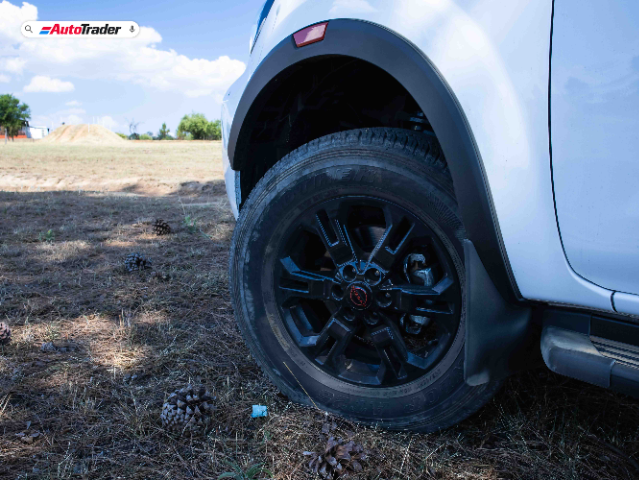 Close-up view of the front right wheel of a white Nissan Navara PRO-4X parked on a grassy field with pinecones scattered around. The image prominently features the black alloy wheel and tire, with the AutoTrader logo visible in the top left corner.