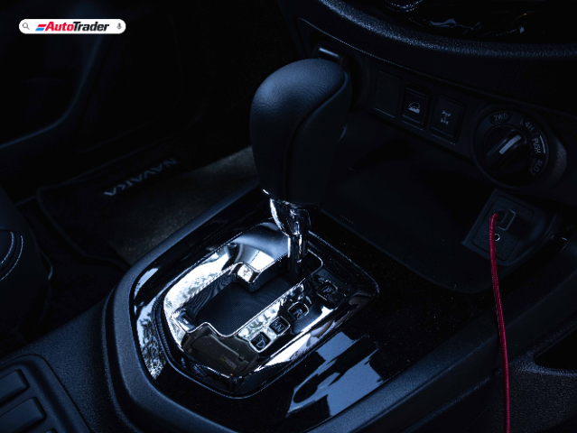The image shows the interior of a Nissan Navara PRO-4X, focusing on the gear shift area. The gear shift lever is prominently displayed in the center, surrounded by a glossy black panel with chrome accents. The gear positions (P, R, N, D) are clearly visible. To the right of the gear shift, there are buttons and a dial for controlling the vehicle's drive modes, including 2WD, 4H, and 4LO. A red cable is plugged into a port near the drive mode controls. The floor mat with the "NAVARA" branding is partially visible in the background. The AutoTrader logo is present at the top left corner of the image.