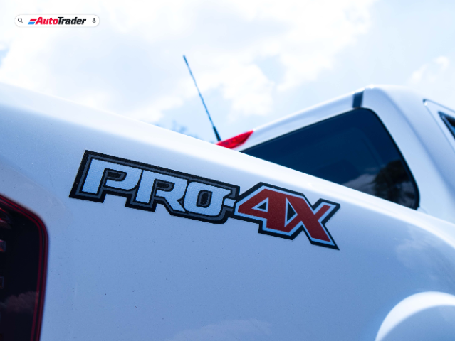 Close-up view of the rear side of a white Nissan Navara PRO-4X, showcasing the PRO-4X badge in black and red. The image is taken from a low angle, highlighting the truck's sleek design under a bright, partly cloudy sky. The AutoTrader logo is visible in the top left corner.