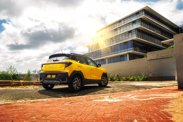 A bright yellow Mahindra XUV3OO is parked on a brick-paved area in front of a modern multi-story glass building. The sun is shining brightly, casting a warm glow on the scene, with a partly cloudy sky in the background. The car is viewed from the rear, showcasing its sleek design and distinctive taillights.