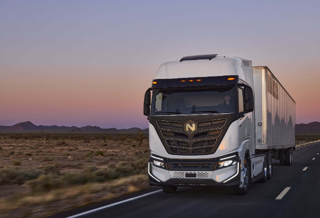 A white semi-truck with a trailer is driving on a desert highway at sunset.