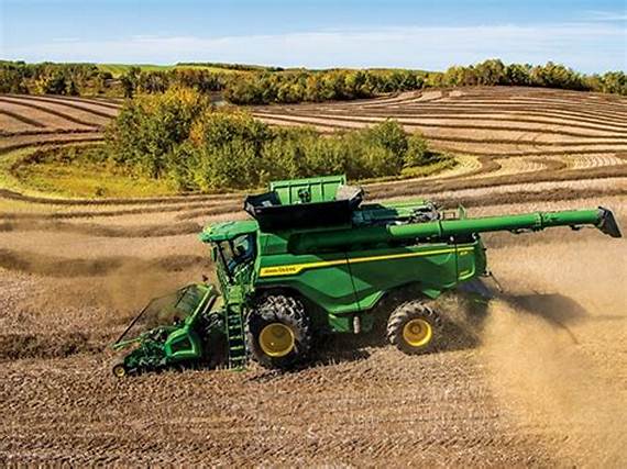 A green combine harvester is working in a large, harvested field with rolling hills and trees in the background.
