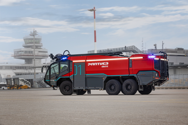 The image shows a red and black electric fire truck labeled "Panther" parked on an airport tarmac with an airport control tower in the background.