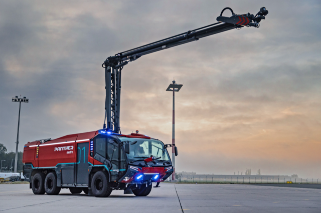 The image shows a large, red, electric fire truck with an extended arm, parked on an airport tarmac at dawn or dusk.
