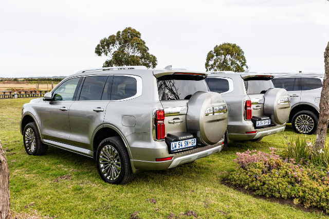 Two silver GWM Tank 500 SUVs are parked on a grassy area. Both vehicles have spare tire covers mounted on their rear doors, and their license plates are visible. The SUVs are positioned side by side, facing away from the camera, with a scenic background of trees and open fields.