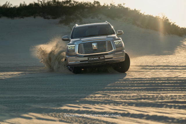 A silver GWM Tank 500 SUV is driving through a sandy desert, kicking up a cloud of sand behind it. The vehicle is shown from the front, with its headlights on and the grille prominently displayed. The sun is setting in the background, casting a warm glow over the scene. A disclaimer at the bottom of the image reads, "Overseas model shown."