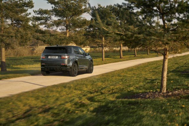 A Land Rover Discovery Sport is driving on a paved road surrounded by green grass and trees. The vehicle is viewed from the rear, showcasing its sleek design and black exterior. The road curves gently, and the scene is set in a serene, natural environment with a clear sky.