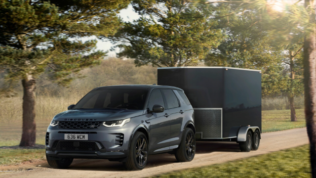 A Land Rover Discovery Sport is towing a large, enclosed trailer on a dirt road surrounded by trees. The vehicle is dark gray with black wheels and tinted windows, and the trailer is also dark in color. The scene is set in a sunny, wooded area with a mix of sunlight and shadows.