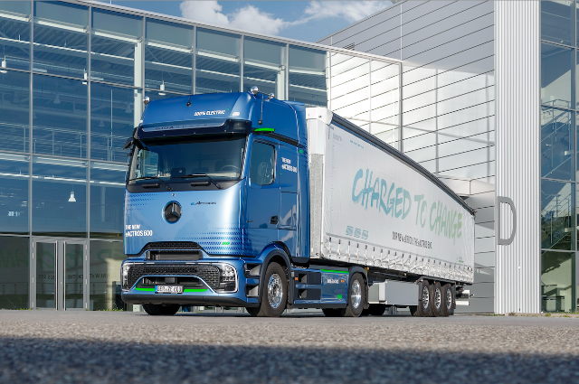 A blue, 100% electric Mercedes-Benz eActros 600 truck is parked in front of a modern glass building, with the slogan "Charged to Change" displayed on the trailer.