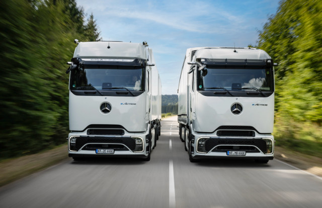 The image shows two white Mercedes-Benz trucks driving side by side on a road surrounded by greenery.