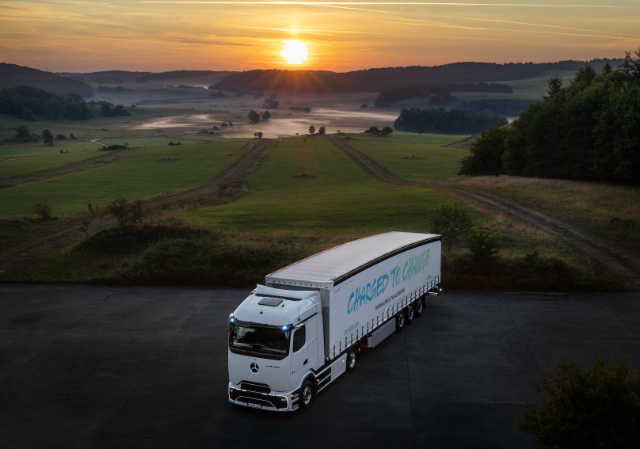 A large white truck labeled "Charged to Change" is parked on a road with a scenic view of a grassy landscape and sunrise in the background.