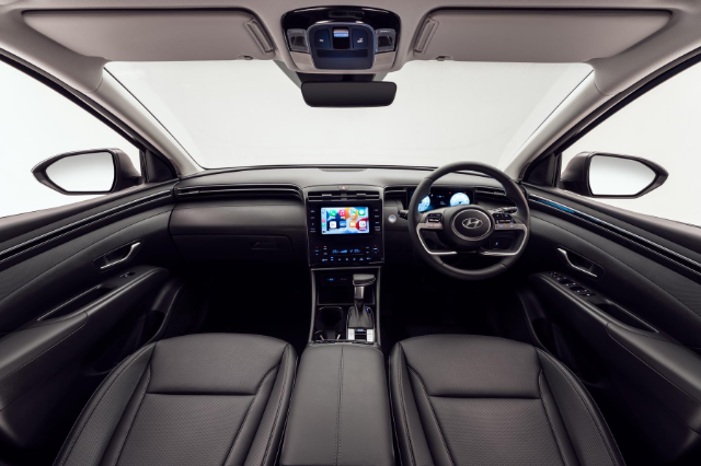 Interior view of a Hyundai Tucson, showcasing the dashboard and steering wheel. The image highlights a modern infotainment system with a touch screen display in the center console. The dashboard features sleek lines and modern design with black leather seats visible in the foreground.